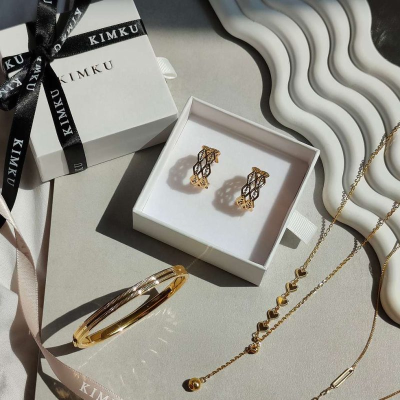 1在陽光照映下的純金手鐲，及純金耳環被收納在專屬有KIMKU品牌緞帶的包裝盒中。